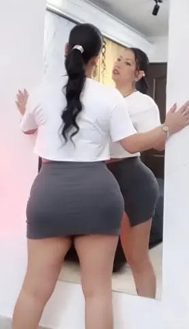 Woman in white t-shirt and grey mini skirt posing in front of a mirror in a bright room.