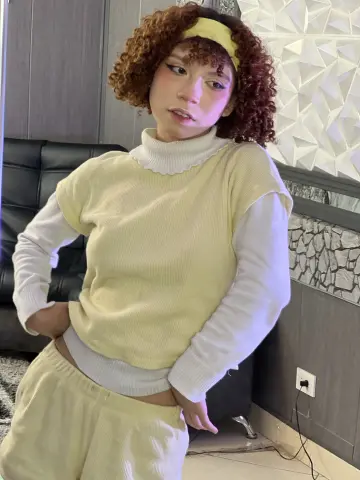 Jessiee_Belle with curly red hair and a yellow headband poses in a pale yellow sweater and matching shorts.