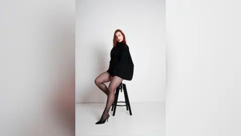 AryaSin poses sitting on a black stool wearing a black long-sleeve shirt and sheer black tights with black high heels against a white background.