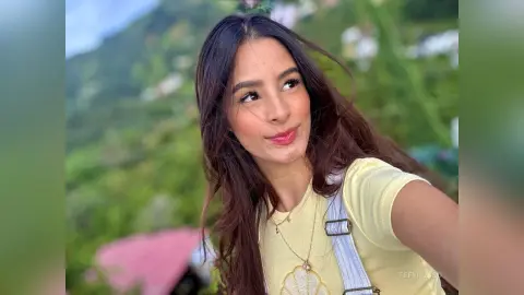 TefyRusso smiles while wearing a yellow t-shirt and light blue denim overalls against a lush, green hillside background.