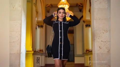 CristalMatthews stands in a grand hallway wearing a tight black long-sleeved mini dress with white trim.