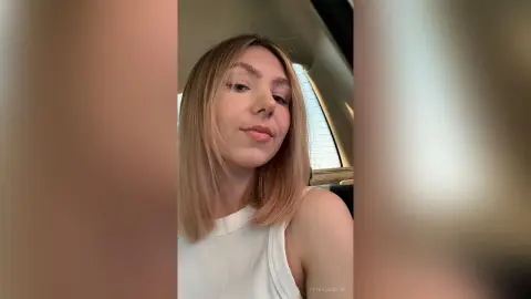 Blonde woman in a white ribbed tank top sitting in a car with a relaxed expression.