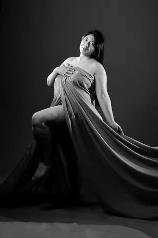 Elegant woman in a flowing draped fabric gown posing gracefully in a moody black and white studio portrait.