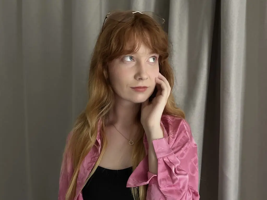 MaryellenQazi has long red hair and light eyes, wearing a pink button-down shirt and a black top while posing against a grey curtain background.
