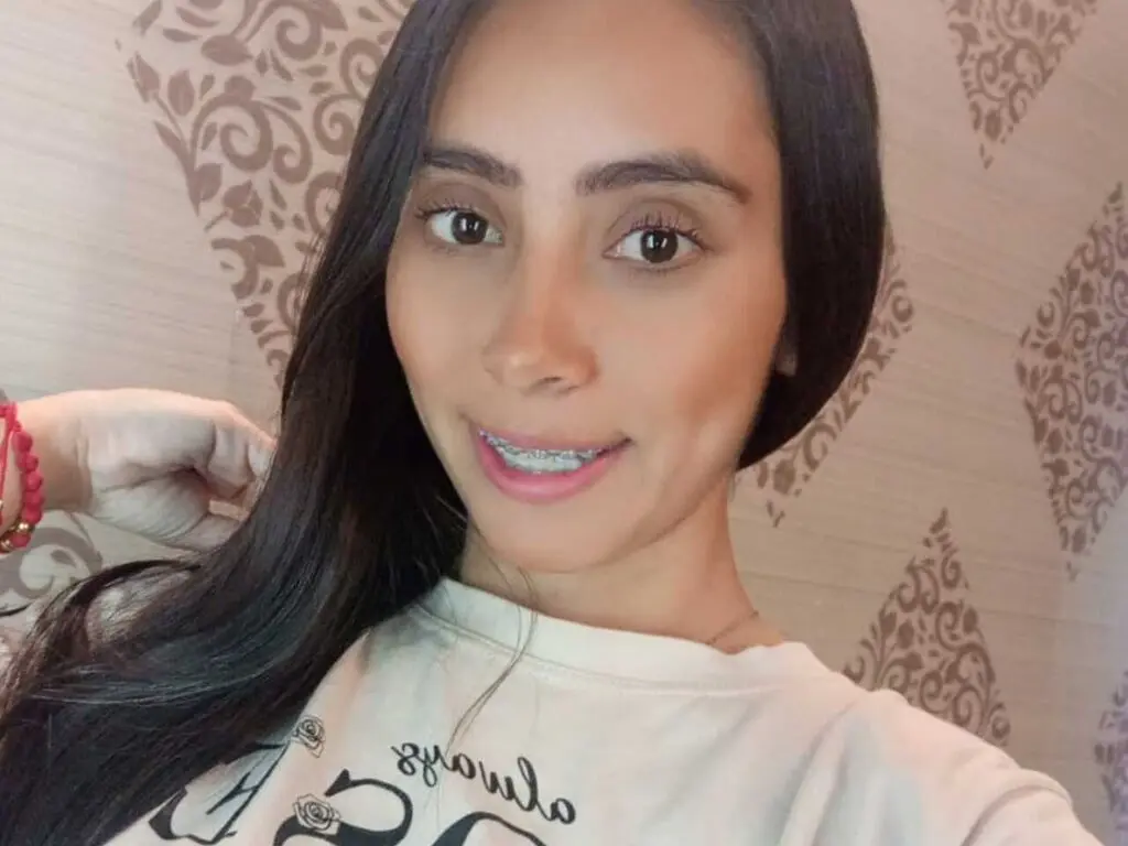 Smiling young woman with long dark hair wearing a white graphic t-shirt in a cozy patterned room.