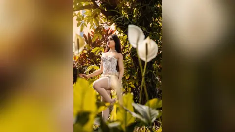 Elegant woman in a white lace corset outfit posing amidst lush green tropical plants in soft sunlight.
