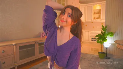 Smiling young woman in a purple ribbed v-neck top posing in a cozy, warm-lit modern living room.