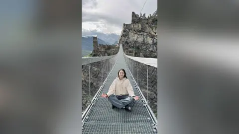 RitaMoonllight sits cross-legged on a metal suspension bridge, wearing a light tan hoodie and grey pants against a backdrop of rocky cliffs and mountains.