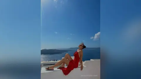 Stylish woman in a red summer dress relaxing on a white balcony overlooking the blue sea in Santorini.