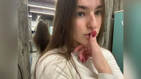 Young woman with long brown hair in a white sweater posing thoughtfully in a modern wood-paneled room.