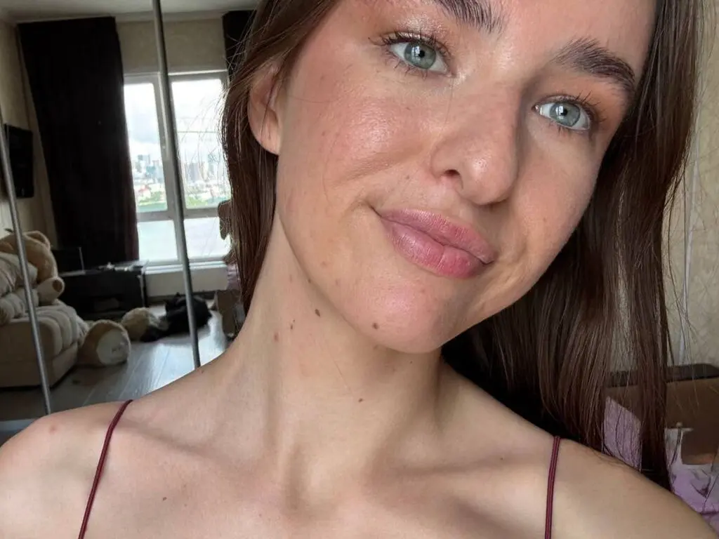 Close-up portrait of a smiling young woman with blue eyes and freckles in a bright indoor setting.