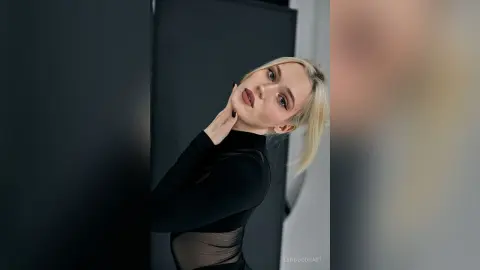 Blonde woman in a black long-sleeve top posing with a moody, elegant expression against a dark background.