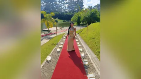Elegant woman in a gold evening gown walking on a red carpet path by a tropical lake at a sunny outdoor event.