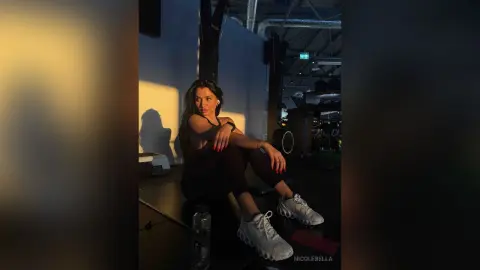 NicoleBella sits on a dark floor wearing black leggings and a black top, posing with one hand on her leg.