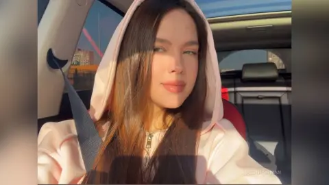 Young woman in a pink hoodie smiling inside a car during a warm golden hour sunset.