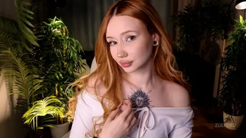 Redheaded woman in a white off-the-shoulder top with a sun chest tattoo sitting near indoor plants.