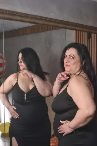Curvy woman in a black sleeveless midi dress posing for a mirror selfie in a modern room.