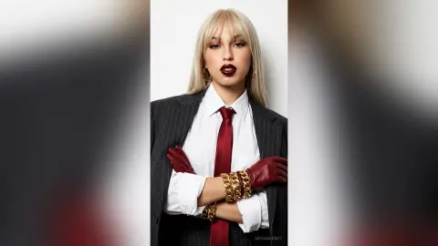 Blonde woman in a black pinstripe suit, white shirt, and red tie wearing burgundy gloves and gold chain jewelry.