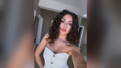 Young woman with curly hair wearing a white strapless top and pearl necklace in a bright indoor setting.