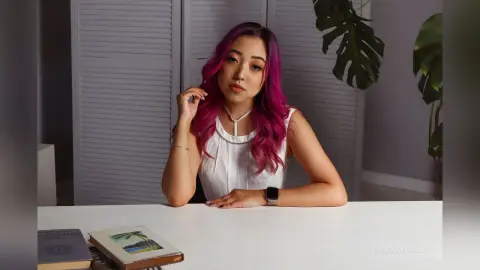 Young woman with vibrant pink hair in a white sleeveless top sitting at a desk in a bright indoor setting.