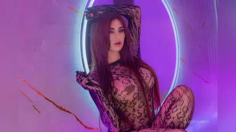 AndreaKalo poses in a long-sleeve snakeskin print bodysuit, sitting in front of a glowing purple ring light with her long brown hair down.