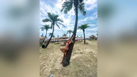 KaylaBryan poses in a red bikini while leaning against a palm tree on a sunny beach.