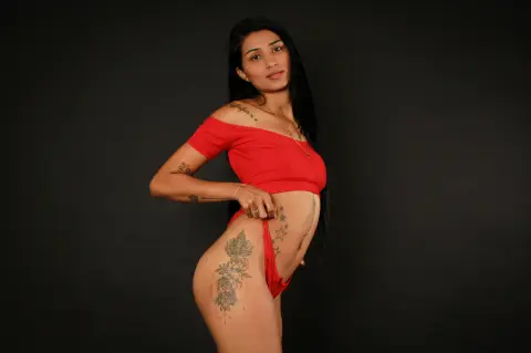 Tattooed woman in a red off-the-shoulder crop top and matching bottoms posing against a black background.