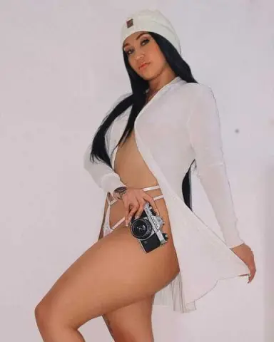 Curvy woman in white knit cardigan and beanie holding a vintage camera against a plain background.