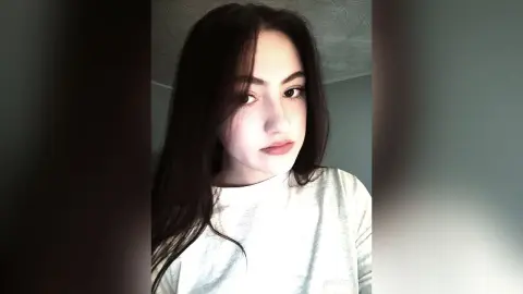 VickyToure poses with long dark hair and a white t-shirt, looking directly at the camera against a plain wall background.