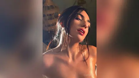 AlessiaMoon poses with wet, dark hair and red lips, showing her bare shoulders and neck in warm lighting.