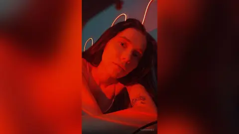 Young woman with long hair in moody red lighting wearing a casual tank top in a dim indoor setting.