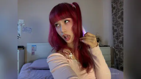 Young woman with long pink hair in a light pink sweater posing playfully in a cozy bedroom setting.