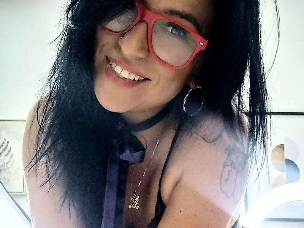 Smiling woman with dark hair and red glasses wearing a black top and gold necklace in bright indoor lighting.