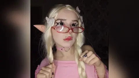Blonde elf cosplay girl with red glasses and pink top posing in a moody indoor setting.