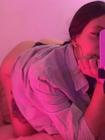 Young woman in a plaid shirt posing in moody pink lighting for a bedroom selfie.