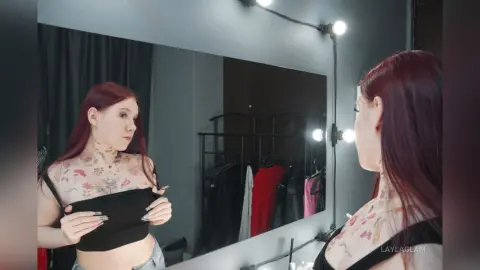 Young woman with red hair and floral tattoos wearing a black crop top in a modern dressing room.