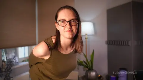 Young woman with glasses in an olive tank top smiling in a cozy, dimly lit indoor setting.