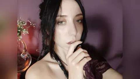 Young woman with dark hair and pale skin wearing a dark lace outfit in a moody, dimly lit setting.