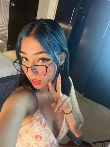 Young woman with blue hair and glasses making a peace sign in a casual indoor setting.