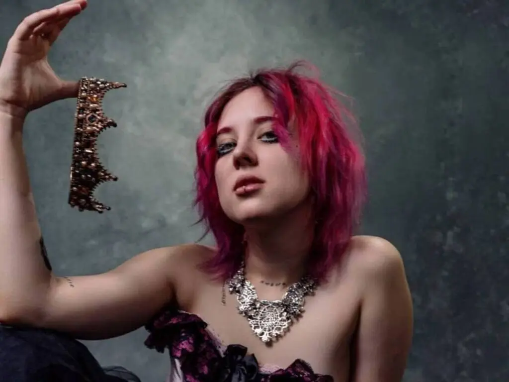 ScarlettGarrett poses with pink hair and a large silver necklace, wearing a dark lace top against a grey background.
