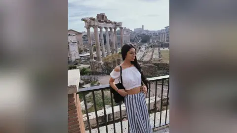 LucyMartin stands on a balcony overlooking ancient stone ruins, wearing a white off-the-shoulder top and blue and white striped trousers.