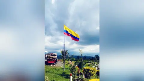 Colombian flag waving on a flagpole against a cloudy sky over a lush green landscape.
