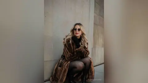 Stylish woman in a brown faux fur coat and black tights posing against a concrete wall in a chic urban setting.
