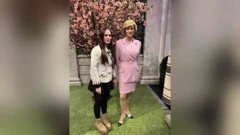 Two smiling women in elegant formal outfits standing before pink cherry blossom trees in a bright garden setting.