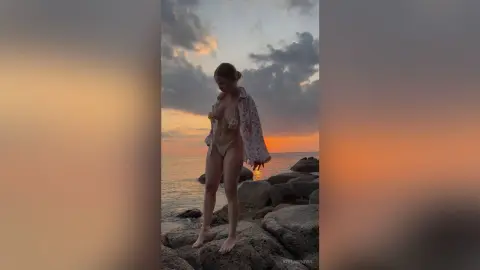 Young woman in a bikini and floral cover-up posing on coastal rocks during a golden sunset.