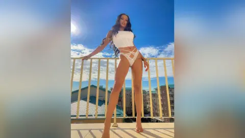 NicoleBella stands on a balcony wearing a white lace corset and matching thong against a bright blue sky.