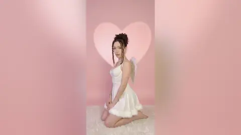Young woman in a white angel costume with wings kneeling against a pink heart background.