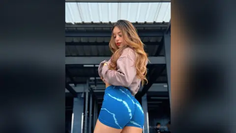 Young woman in blue athletic shorts and a mauve sweatshirt posing confidently in a modern industrial setting.
