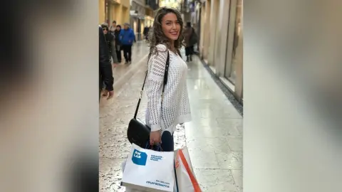 MontseFerrera stands on a city sidewalk wearing a white lace long-sleeve top and carrying a black shoulder bag and shopping bags.