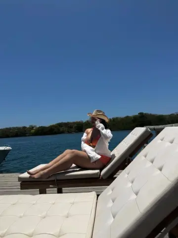 Relaxed woman in an orange bikini and white shirt lounging on a sunbed by a calm blue lake under a clear sky.